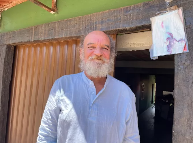 A imagem mostra José Eustáquio em frente a uma casa verde com portão marrom. José é um homem branco e tem barba branca. Ele está usando uma camisa branca de manga comprida e sorrindo para a foto.