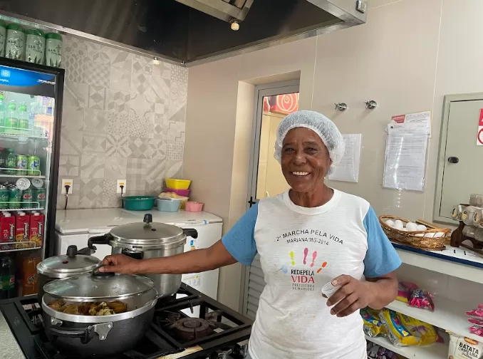  Imagem de Darsila das Graças cozinhando na cozinha de seu restaurante. Darsila está no fogão abrindo a tampa da panela, olhando para a câmera sorrindo. Ela usa uma touca na cabeça e blusa branca. Ao fundo uma geladeira com bebidas e ao lado um freezer.