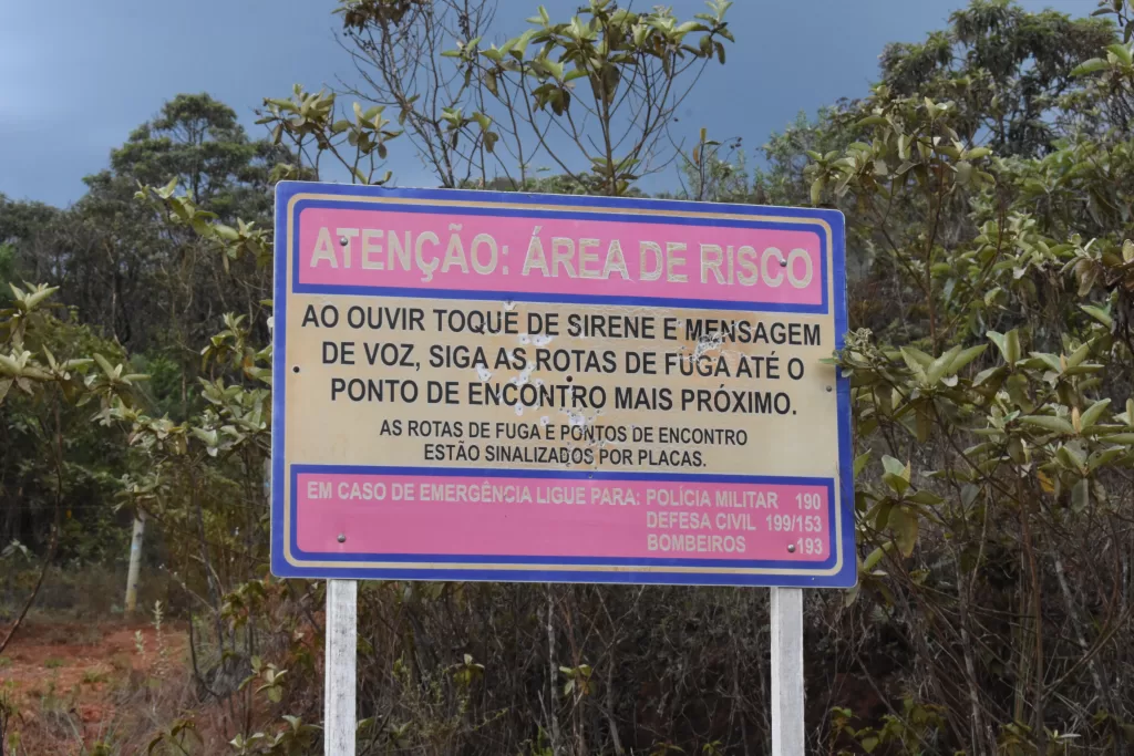 A imagem mostra uma placa bege, com bordas azuis, e dois espaços rosa dentro. O primeiro espaço está escrito de bege, em caixa alta: ATENÇÃO ÁREA DE RISCO. Na parte de fundo bege da placa, está escrito de preto e com uma letra grande também em caixa alta: AO OUVIR TOQUE DE SIRENE E MENSAGEM DE VOZ, SIGA AS ROTAS DE FUGA ATÉ O PONTO DE ENCONTRO MAIS PRÓXIMO. Abaixo, ainda no fundo bege, está escrito com letras pretas, caixa alta, mas menores: AS ROTAS DE FUGA E PONTOS DE ENCONTRO ESTÃO SINALIZADOS POR PLACAS. No segundo espaço rosa, logo abaixo do fundo bege, está escrito com letras bege: EM CASO DE EMERGÊNCIA LIGUE PARA: POLICIA MILITAR 190, DEFESA CIVIL 199/153, BOMBEIROS 193. Atrás da placa tem algumas árvores, acima das árvores um céu azul, e abaixo da placa, há uma vegetação rasteira, mostrando um pouco de terra.