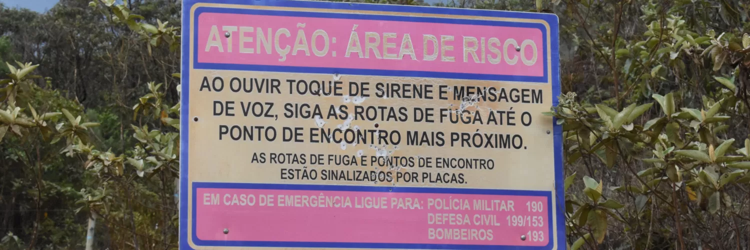 A imagem mostra uma placa bege, com bordas azuis, e dois espaços rosa dentro. O primeiro espaço está escrito de bege, em caixa alta: ATENÇÃO ÁREA DE RISCO. Na parte de fundo bege da placa, está escrito de preto e com uma letra grande também em caixa alta: AO OUVIR TOQUE DE SIRENE E MENSAGEM DE VOZ, SIGA AS ROTAS DE FUGA ATÉ O PONTO DE ENCONTRO MAIS PRÓXIMO. Abaixo, ainda no fundo bege, está escrito com letras pretas, caixa alta, mas menores: AS ROTAS DE FUGA E PONTOS DE ENCONTRO ESTÃO SINALIZADOS POR PLACAS. No segundo espaço rosa, logo abaixo do fundo bege, está escrito com letras bege: EM CASO DE EMERGÊNCIA LIGUE PARA: POLICIA MILITAR 190, DEFESA CIVIL 199/153, BOMBEIROS 193. Atrás da placa tem algumas árvores, acima das árvores um céu azul, e abaixo da placa, há uma vegetação rasteira, mostrando um pouco de terra.