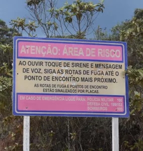 A imagem mostra uma placa bege, com bordas azuis, e dois espaços rosa dentro. O primeiro espaço está escrito de bege, em caixa alta: ATENÇÃO ÁREA DE RISCO. Na parte de fundo bege da placa, está escrito de preto e com uma letra grande também em caixa alta: AO OUVIR TOQUE DE SIRENE E MENSAGEM DE VOZ, SIGA AS ROTAS DE FUGA ATÉ O PONTO DE ENCONTRO MAIS PRÓXIMO. Abaixo, ainda no fundo bege, está escrito com letras pretas, caixa alta, mas menores: AS ROTAS DE FUGA E PONTOS DE ENCONTRO ESTÃO SINALIZADOS POR PLACAS. No segundo espaço rosa, logo abaixo do fundo bege, está escrito com letras bege: EM CASO DE EMERGÊNCIA LIGUE PARA: POLICIA MILITAR 190, DEFESA CIVIL 199/153, BOMBEIROS 193. Atrás da placa tem algumas árvores, acima das árvores um céu azul, e abaixo da placa, há uma vegetação rasteira, mostrando um pouco de terra.