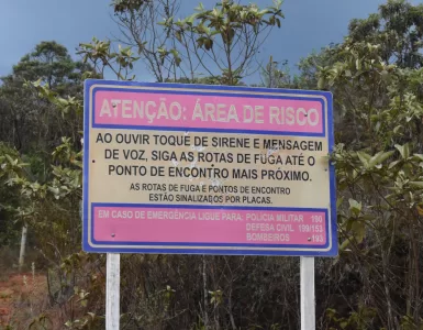 A imagem mostra uma placa bege, com bordas azuis, e dois espaços rosa dentro. O primeiro espaço está escrito de bege, em caixa alta: ATENÇÃO ÁREA DE RISCO. Na parte de fundo bege da placa, está escrito de preto e com uma letra grande também em caixa alta: AO OUVIR TOQUE DE SIRENE E MENSAGEM DE VOZ, SIGA AS ROTAS DE FUGA ATÉ O PONTO DE ENCONTRO MAIS PRÓXIMO. Abaixo, ainda no fundo bege, está escrito com letras pretas, caixa alta, mas menores: AS ROTAS DE FUGA E PONTOS DE ENCONTRO ESTÃO SINALIZADOS POR PLACAS. No segundo espaço rosa, logo abaixo do fundo bege, está escrito com letras bege: EM CASO DE EMERGÊNCIA LIGUE PARA: POLICIA MILITAR 190, DEFESA CIVIL 199/153, BOMBEIROS 193. Atrás da placa tem algumas árvores, acima das árvores um céu azul, e abaixo da placa, há uma vegetação rasteira, mostrando um pouco de terra.