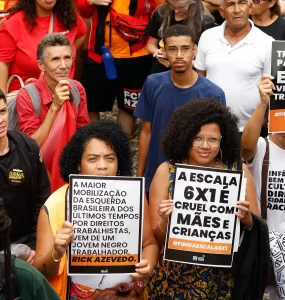 Duas mulheres negras em uma manifestação segurando cartazes com frases como “A escala 6x1 é cruel com mães e crianças” e “Trabalhar para viver, não viver para trabalhar”. O protesto ocorreu em um ambiente público com várias pessoas ao fundo, algumas usando camisetas e bandeiras vermelhas.