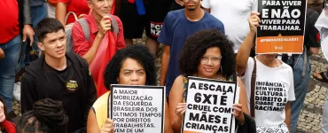 Duas mulheres negras em uma manifestação segurando cartazes com frases como “A escala 6x1 é cruel com mães e crianças” e “Trabalhar para viver, não viver para trabalhar”. O protesto ocorreu em um ambiente público com várias pessoas ao fundo, algumas usando camisetas e bandeiras vermelhas.