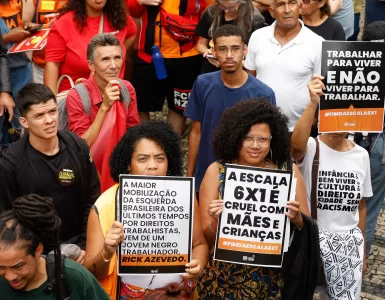 Duas mulheres negras em uma manifestação segurando cartazes com frases como “A escala 6x1 é cruel com mães e crianças” e “Trabalhar para viver, não viver para trabalhar”. O protesto ocorreu em um ambiente público com várias pessoas ao fundo, algumas usando camisetas e bandeiras vermelhas.