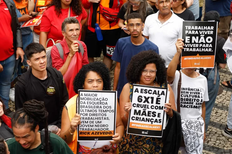 Duas mulheres negras em uma manifestação segurando cartazes com frases como “A escala 6x1 é cruel com mães e crianças” e “Trabalhar para viver, não viver para trabalhar”. O protesto ocorreu em um ambiente público com várias pessoas ao fundo, algumas usando camisetas e bandeiras vermelhas.