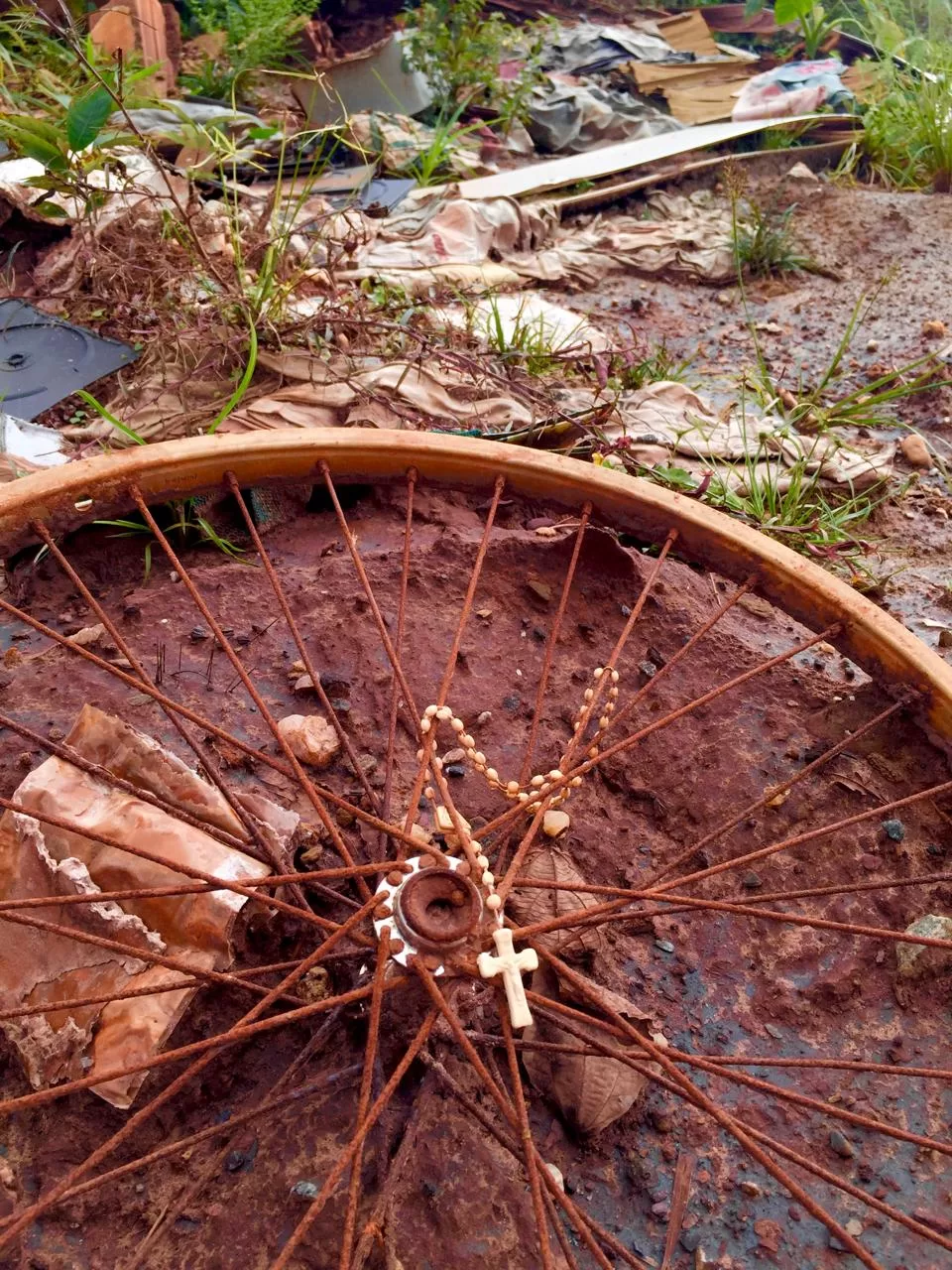 A imagem mostra um terço preso a uma roda enferrujada, em meio a barro e destroços.