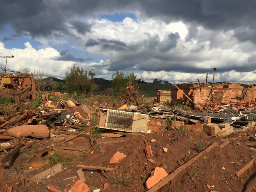 A foto mostra uma região com muitos destroços, sendo possível ver uma casa sem o telhado ao fundo, uma geladeira caída no centro, tijolo e madeira espalhados também.