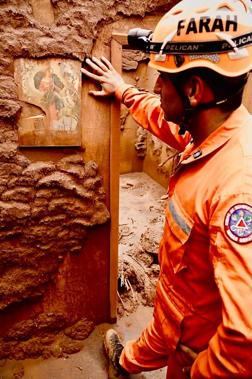  Foto do bombeiro Leo Farah observando uma pintura em uma casa totalmente tomada pela lama. Leo usa o uniforme e o capacete dos bombeiros laranja 
inserir foto 8
