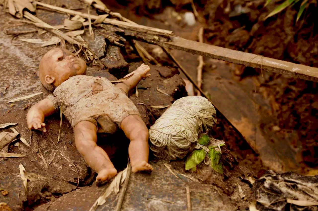 A fotografia mostra uma boneca suja de barro e um novelo de lã em cima de escombros, e ao lado, destroços de madeira.