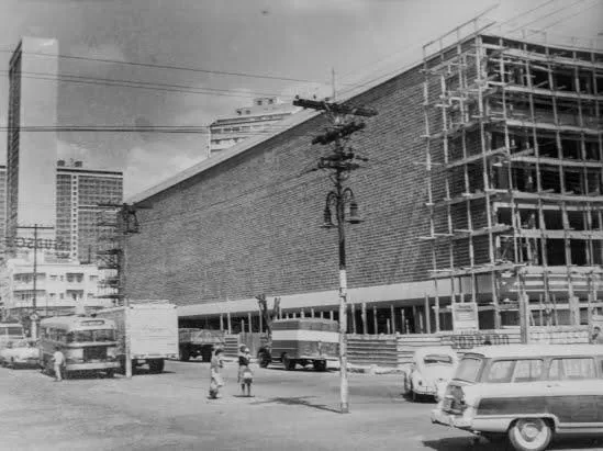 Imagem em preto e branco da década de 60. Mostra a construção em andamento, quase finalizada, do edifício do Mercado Novo. O prédio tem paredes de tijolos estilo cobogó e andaimes expostos no lado direito. Na rua, há movimento de veículos, incluindo ônibus, caminhões e carros antigos. Também é possível ver pessoas caminhando, postes de iluminação e fiação elétrica ao longo da via. No fundo, aparecem alguns prédios.