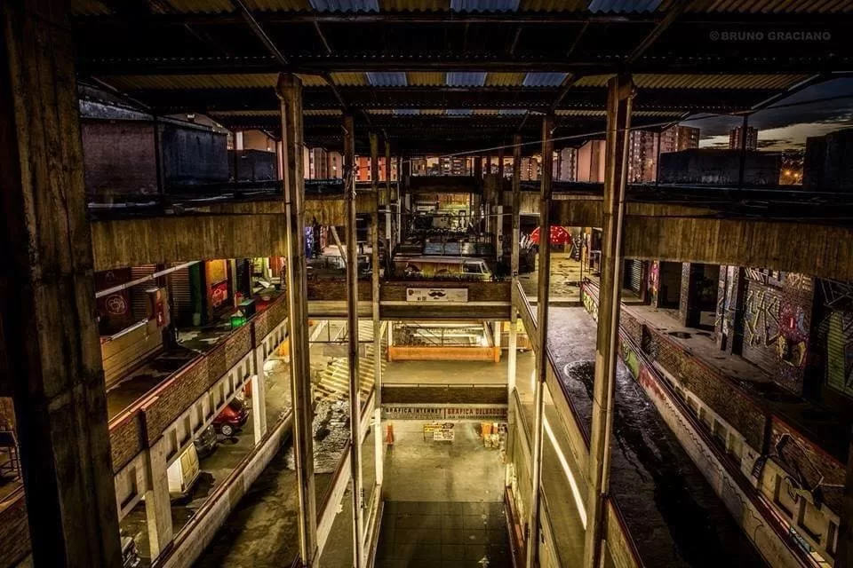 A imagem retrata o interior do Mercado Novo antes de sua revitalização, com um ambiente marcado por estruturas de concreto aparente e um teto metálico. O espaço possui vários andares interligados por passarelas e escadas, com iluminação amarela e baixa. As paredes estão cobertas por grafites e marcas de desgaste, evidenciando o estado de abandono do local na época. No térreo, há áreas abertas e veículos estacionados no segundo piso. Ao fundo, é possível observar prédios contemplados pela iluminação do fim de tarde.