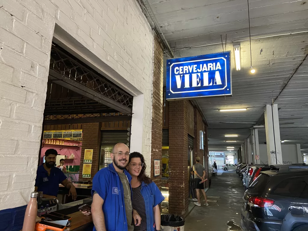 Lateral da loja Cervejaria Viela com tijolos pintados de branco e azul, acima há uma placa azul com letras brancas. Há um balcão onde está o gerente do turno Gabriel Dabian ao lado de uma funcionária, ambos usam uniforme azul, óculos e possuem pele clara. Ao fundo há outro funcionário sentado de uniforme azul, barba e cabelos escuros. O ambiente mostra outras lojas do mercado e um trecho do estacionamento.