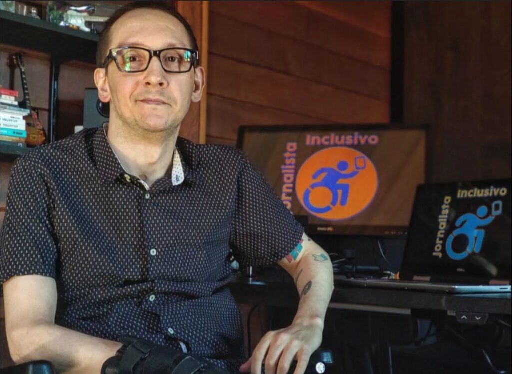 Homem Branco usando blusa social azul, óculos de grau com armação preta, posando sentado. Ao fundo, livros e monitores de computador com a logomarca do Jornalista Inclusivo.