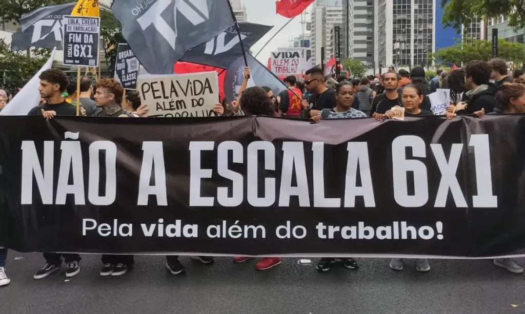 Um grupo de manifestantes segura uma grande faixa preta com os dizeres “NÃO À ESCALA 6x1 - Pela vida além do trabalho!”, enquanto outras pessoas ao fundo exibem cartazes com mensagens de protesto. A manifestação ocorre em um cenário urbano, com prédios ao redor.
