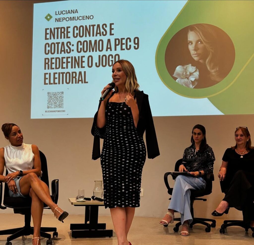 Mulher branca de vestido preto de tubinho com bolinhas brancas em um palco falando no microfone com algumas pessoas dispostas atrás.