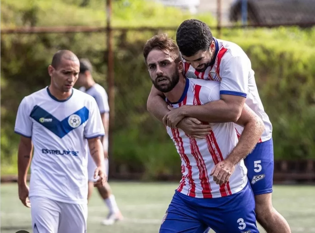 Dois jogadores de camisa vermelha e branca se abraçando comemorando um gol. Ao fundo, o atleta rival lamenta o acontecido