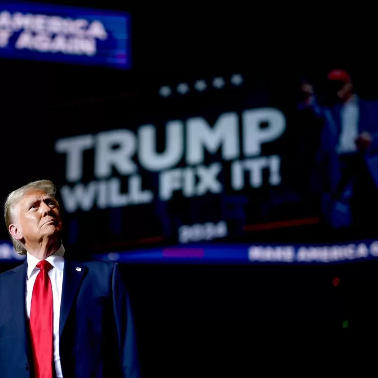 O presidente eleito dos Estados Unidos, Donald Trump, de pé, usando terno cor azul marinho e gravata vermelha, em frente a um telão escrito “Trump Will Fix It” (“Trump irá consertar tudo”, traduzido do inglês).