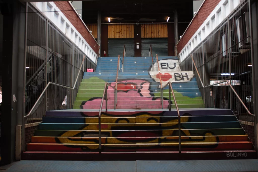 Escadaria grafitada, colorida com a arte de um bolinho amarelo com cobertura rosa e uma cereja. Ao lado do bolinho, há um balão de fala escrito “EU AMO BH”.
