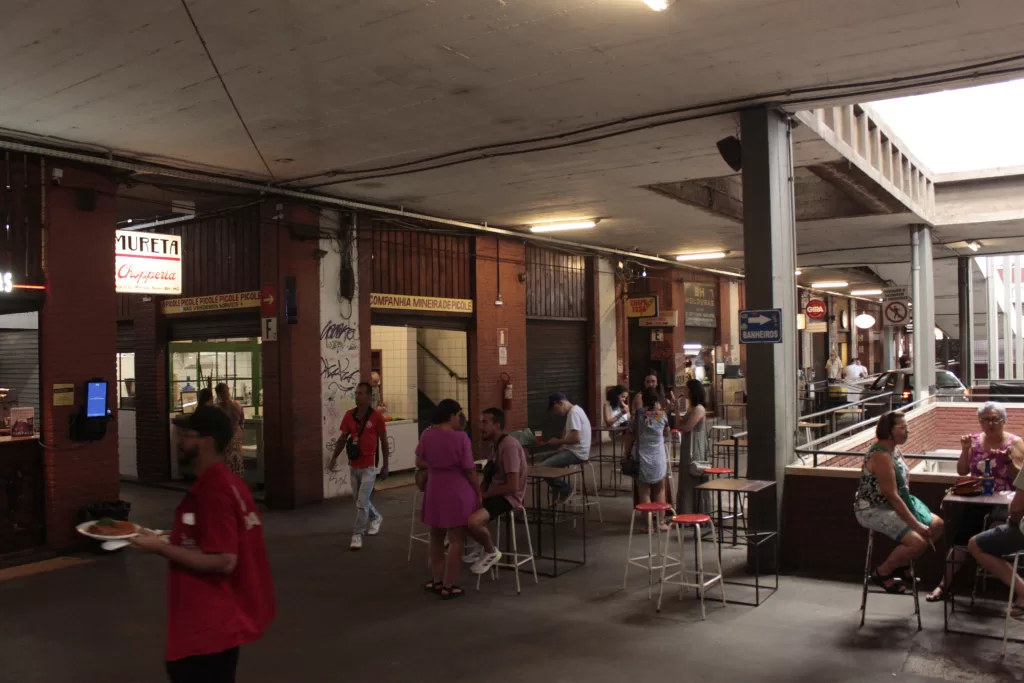 Imagem com uma visão ampla do mercado durante o dia. No canto superior esquerdo há uma placa com fundo branco e caracteres pretos e vermelhos escrito “MURETA CHOPPERIA”. Abaixo, no canto inferior esquerdo, um homem de camisa vermelha e boné andando com um prato de comida nas mãos. Ao centro da foto, há algumas pessoas sentadas em bancos e mesinhas. Ao fundo da imagem, uma loja com a placa em amarelo e caracteres vermelhos escrito “COMPANHIA MINEIRA DE PICOLÉ”.

