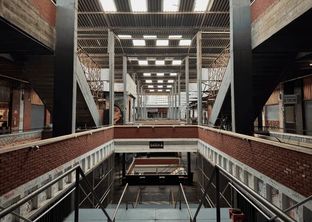 A imagem mostra o interior do Mercado com arquitetura industrial. Há estruturas de concreto e metal, com paredes de tijolos e um telhado elevado com painéis transparentes que possibilitam a entrada de luz natural. No centro da imagem, uma placa verde indica “SAÍDA” com uma seta apontando para baixo. Escadas conectam o andar superior ao inferior, onde é possível ver mais áreas de circulação e algumas lojas fechadas. No nível superior, há murais artísticos nas paredes.