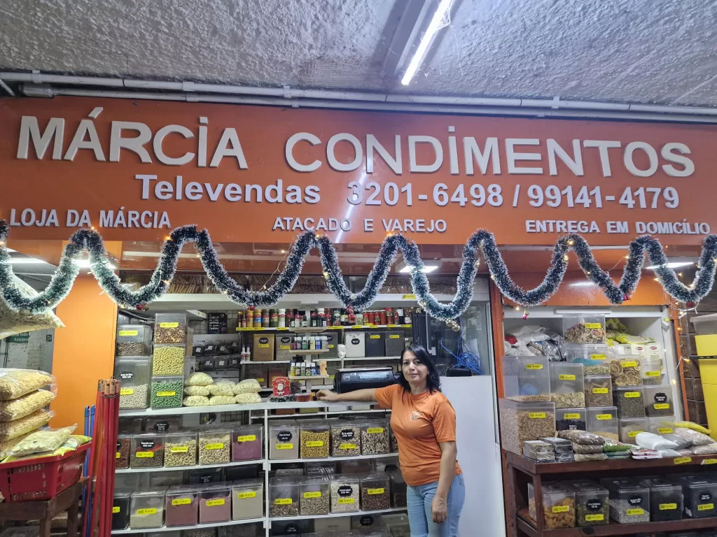 Loja “Márcia Condimentos”, fachada de cor laranja e letras brancas com decoração de natal. Em frente à loja, há uma mulher de camiseta laranja e calça jeans. Pele clara, cabelo preto, liso e curto, olhos pretos e sorri. O interior da loja conta com prateleiras e recipientes transparentes com diversos tipos de condimentos.
