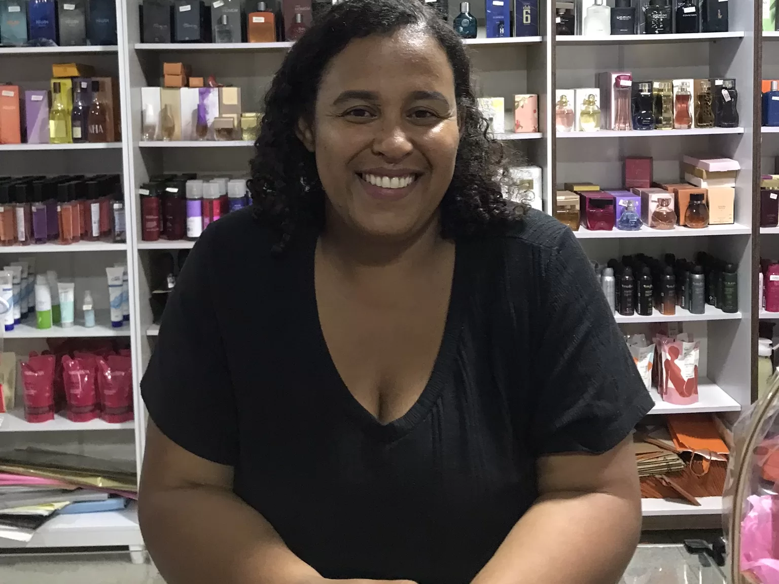 A imagem mostra a empreendedora Célia Maria, com uma blusa preta, sorrindo para a câmera. Atrás estão expostos cosméticos de diferentes tipos.