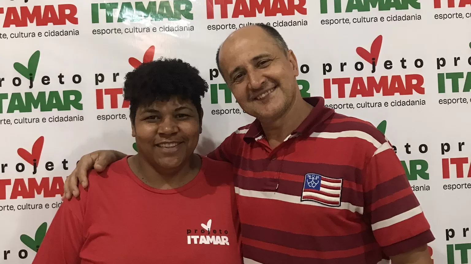 Os empreendedores Fernanda Benevides e Itamar José da Silva estão sorrindo para a foto, Itamar está com a mão no ombro de Fernanda. Ela veste uma blusa vermelha com a logomarca do projeto, Itamar está com uma blusa vermelha com listras horizontais em vinho e vermelho. O painel atrás contém a logomarca do Projeto Itamar.