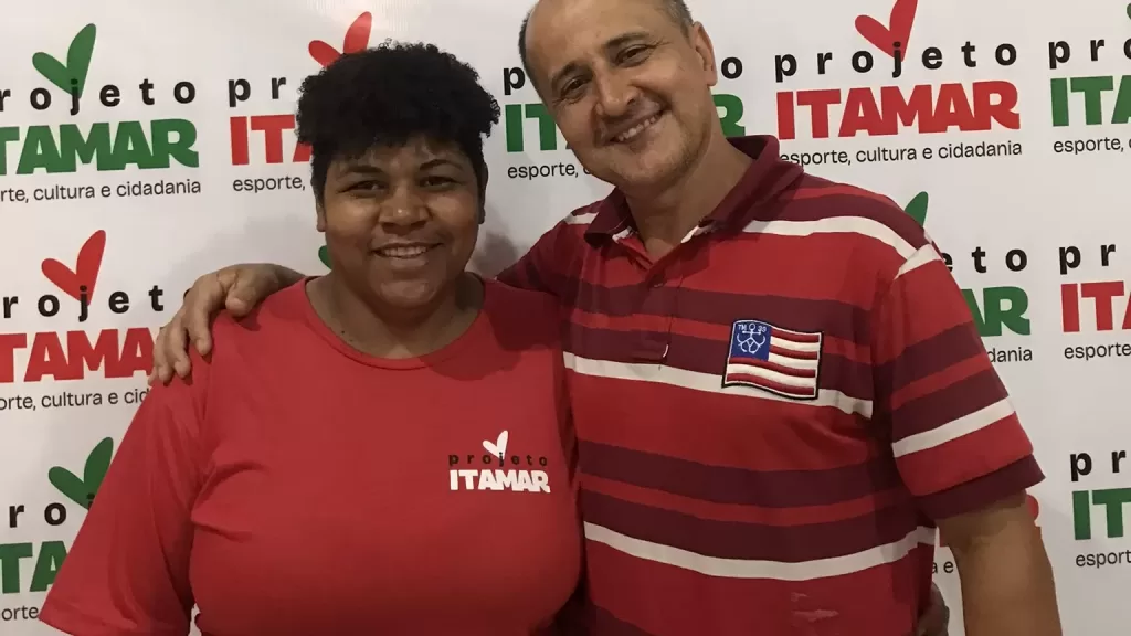 Os empreendedores Fernanda Benevides e Itamar José da Silva estão sorrindo para a foto, Itamar está com a mão no ombro de Fernanda. Ela veste uma blusa vermelha com a logomarca do projeto, Itamar está com uma blusa vermelha com listras horizontais em vinho e vermelho. O painel atrás contém a logomarca do Projeto Itamar.