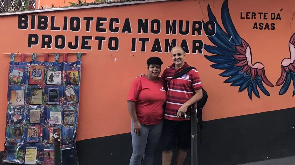 Os empreendedores Fernanda e Itamar em frente a um muro laranja. No centro do muro está escrito "Biblioteca no muro. Projeto Itamar" e ao lado "Ler te dá asas", com asas coloridas em baixo. No muro, está pendurado um painel de tecido com livros.