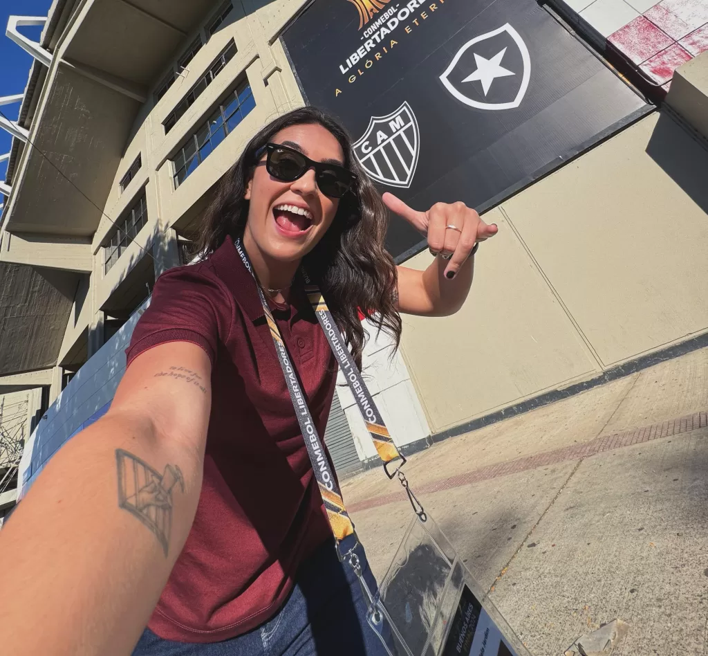Mulher branca, vestida com uma blusa vermelha e calça, e usando óculos escuro, do lado de fora do estádio. Ela sorri e faz gesto de relaxamento com a mão. Ao fundo, um template com os símbolos de Atlético-MG e Botafogo-RJ movimentado.