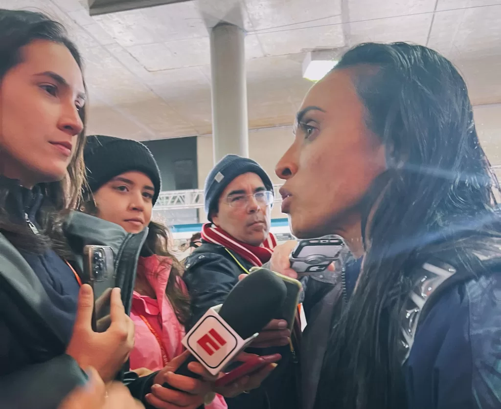 Mariana, mulher branca de roupa preta, segura o microfone da ESPN em uma mão e na outra segura um celular enquanto entrevista a jogadora Marta, que veste o uniforme da Seleção Brasileira. As duas estão em pé, próximas uma da outra, com semblante sério e atento.