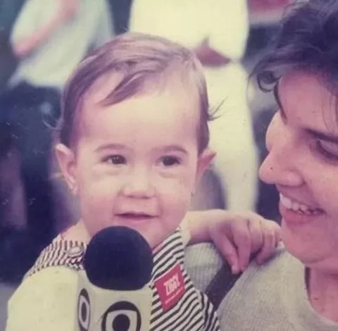 Adriana Spinelli, uma mulher adulta com uma criança no colo sorrindo, sua filha Mariana Spinelli. A criança também está sorrindo e segura um microfone