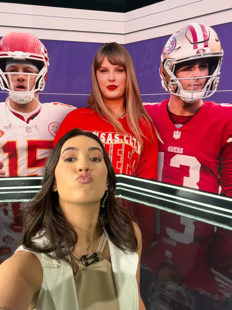 Mulher branca de cabelos castanhos tirando uma foto do seu próprio rosto. Mariana Spinelli está dentro de um estúdio de gravação. Atrás dela há um telão que está com imagem fixada de dois homens jogadores de futebol americano vestidos com camisas branca e vermelha e capacetes branco e vermelho. No meio da imagem, entre os dois uma mulher loira de roupa vermelha.