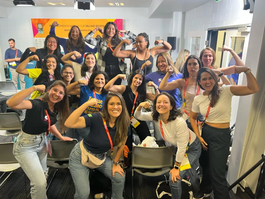 Grupo de cerca de quinze mulheres sorrindo e posando para foto em uma sala destinada à imprensa. O grupo aparenta estar animado e celebrando um momento especial durante um evento esportivo internacional.