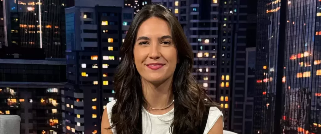 Mulher branca de cabelos castanhos sentada de frente para a câmera e sorrindo no estúdio do SportCenter com uma imagem de prédios ao fundo. Veste Blusa branca com um colete preto, calça marrom