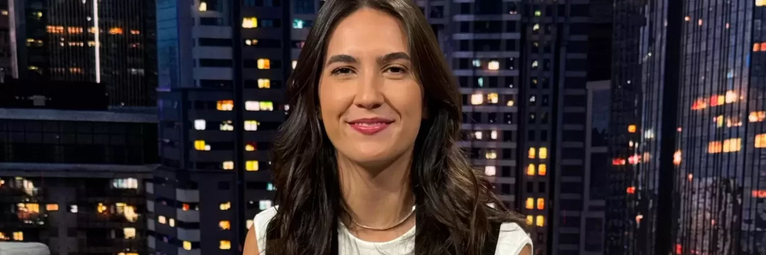 Mulher branca de cabelos castanhos sentada de frente para a câmera e sorrindo no estúdio do SportCenter com uma imagem de prédios ao fundo. Veste Blusa branca com um colete preto, calça marrom