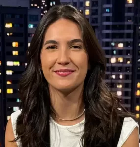 Mulher branca de cabelos castanhos sentada de frente para a câmera e sorrindo no estúdio do SportCenter com uma imagem de prédios ao fundo. Veste Blusa branca com um colete preto, calça marrom