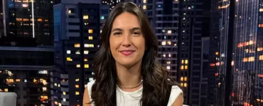 Mulher branca de cabelos castanhos sentada de frente para a câmera e sorrindo no estúdio do SportCenter com uma imagem de prédios ao fundo. Veste Blusa branca com um colete preto, calça marrom