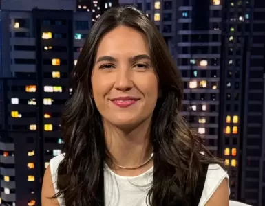 Mulher branca de cabelos castanhos sentada de frente para a câmera e sorrindo no estúdio do SportCenter com uma imagem de prédios ao fundo. Veste Blusa branca com um colete preto, calça marrom