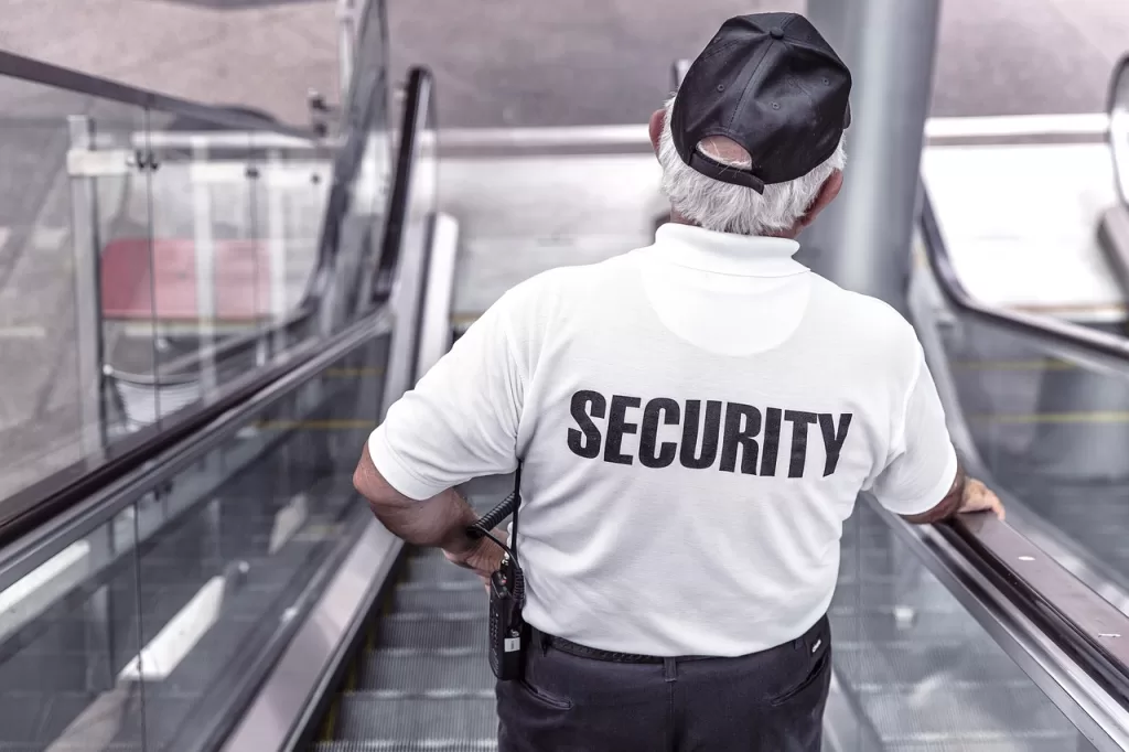 Homem com cabelos brancos e um boné na cabeça, utilizando uma camisa escrito "security" (segurança,em português)