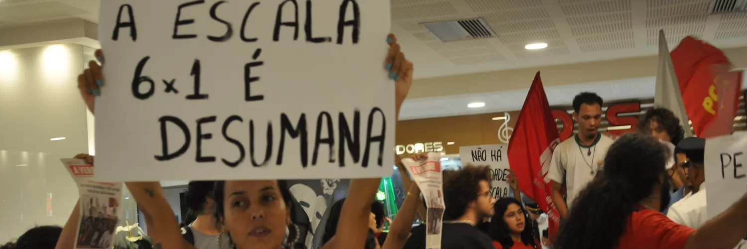 Foto de uma manifestação a favor do fim da jornada 6x1. Há uma mulher branca de cabelo escuro que segura uma placa escrira "a escala 6x1 é desumana" no centro-esquerda da foto
