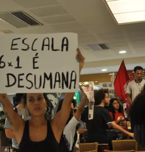 Foto de uma manifestação a favor do fim da jornada 6x1. Há uma mulher branca de cabelo escuro que segura uma placa escrira "a escala 6x1 é desumana" no centro-esquerda da foto
