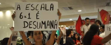 Foto de uma manifestação a favor do fim da jornada 6x1. Há uma mulher branca de cabelo escuro que segura uma placa escrira "a escala 6x1 é desumana" no centro-esquerda da foto