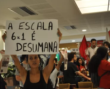 Foto de uma manifestação a favor do fim da jornada 6x1. Há uma mulher branca de cabelo escuro que segura uma placa escrira "a escala 6x1 é desumana" no centro-esquerda da foto