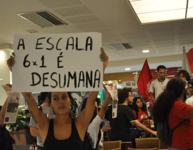 Foto de uma manifestação a favor do fim da jornada 6x1. Há uma mulher branca de cabelo escuro que segura uma placa escrira "a escala 6x1 é desumana" no centro-esquerda da foto