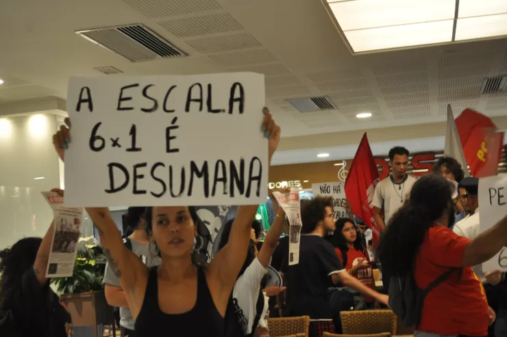 Foto de uma manifestação a favor do fim da jornada 6x1. Há uma mulher branca de cabelo escuro que segura uma placa escrira "a escala 6x1 é desumana" no centro-esquerda da foto