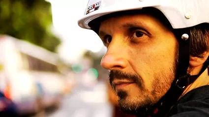 Homem de perfil com barba e capacete branco aparece em close, com expressão séria, olhando para a direita em um cenário urbano desfocado ao fundo, sugerindo que está prestes a se locomover.
