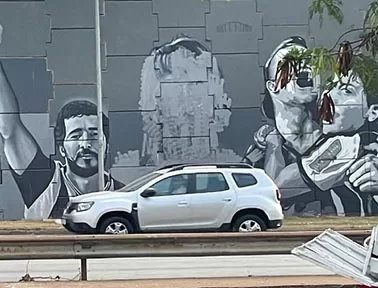 Fotografia de um mural pintado em tons de cinza na parede externa de uma rodovia. A arte retrata três figuras humanas: à esquerda, um homem com o braço erguido em sinal de comemoração; à direita, duas pessoas olhando para cima, com expressões de admiração, e ao centro, Fred Melo Paiva apagado.