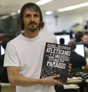 Com uma redação de jornal ao fundo e desfocada, Fred aparece em primeiro plano, de camiseta branca, cabelos castanhos lisos, na altura do queixo, bigode e barba rala, segurando o livro de sua autoria que tem a capa preta e branca, nas cores do Atlético.